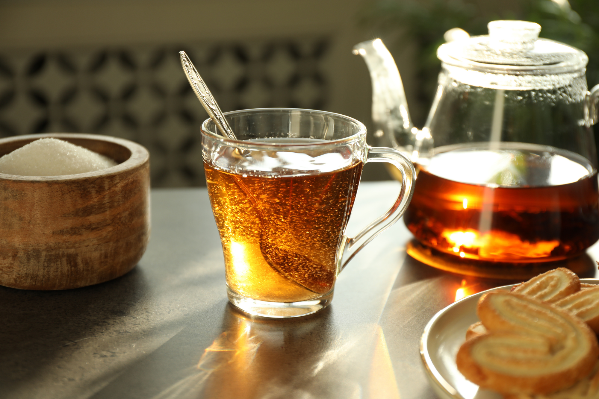 A close-up of a large spoon stirring granulated sugar into the hot, dark tea in the glass.