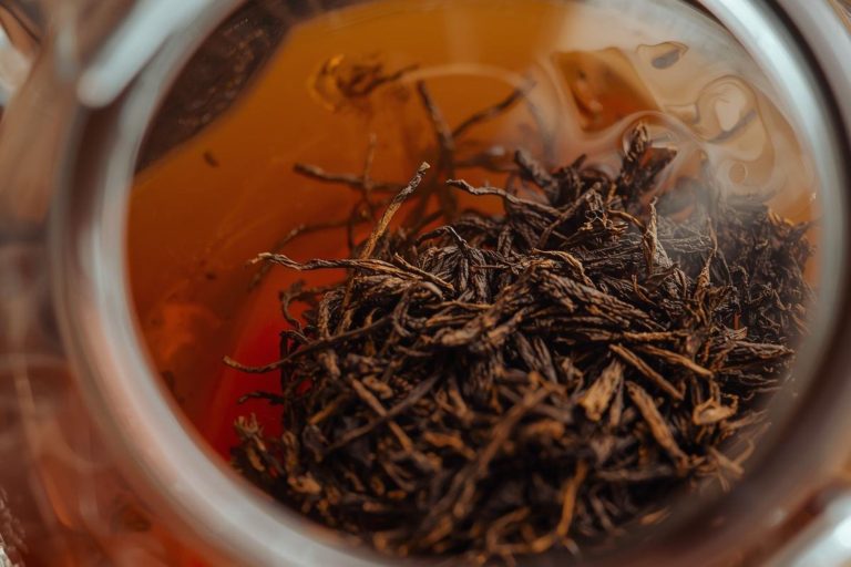 A cozy close-up photo showing loose-leaf hojicha steeping in a clear glass teapot, emphasizing the rich, warm, amber/brown color of the brew.